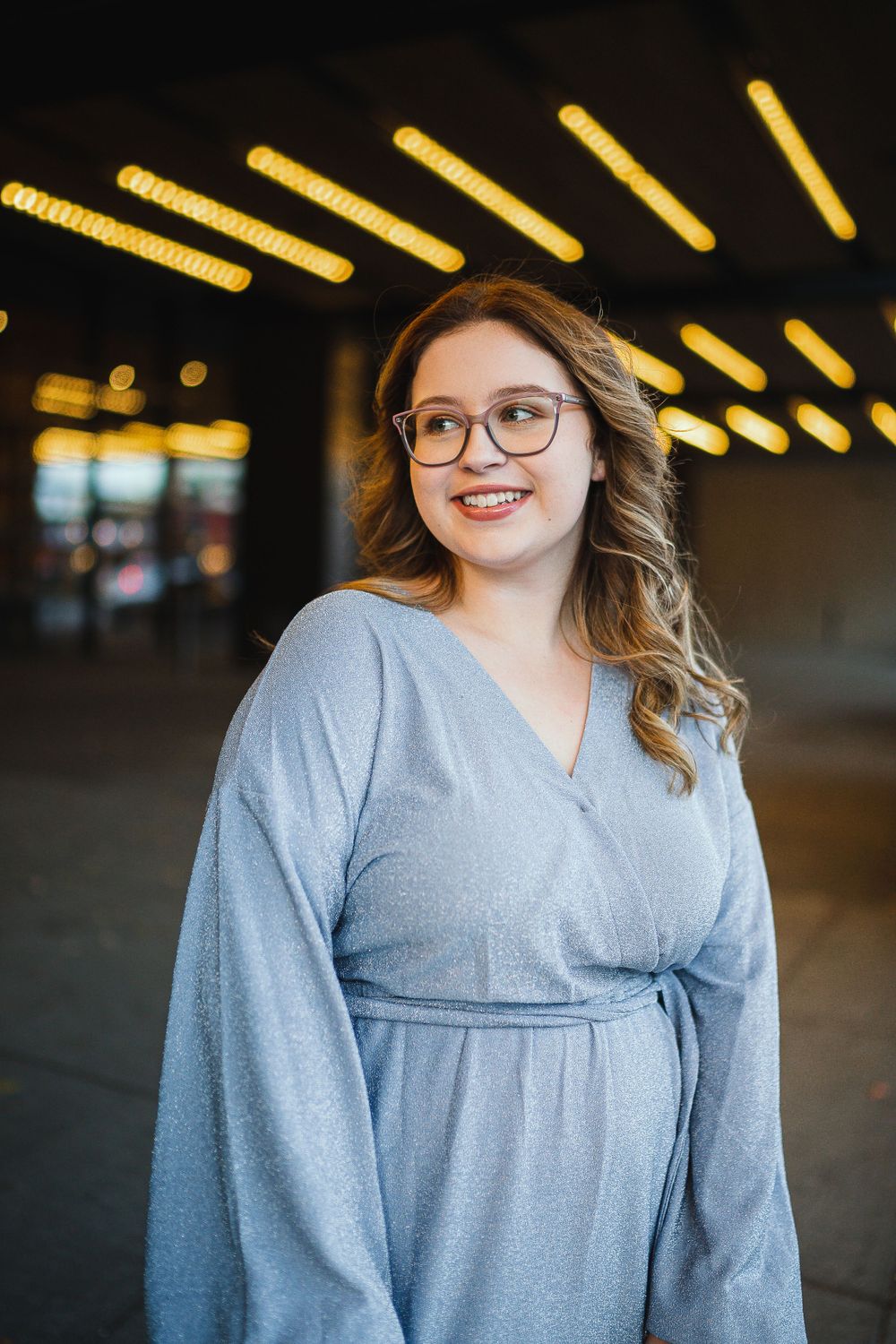 Portrait under yellow lighting wearing blue wrap dress and glasses.
