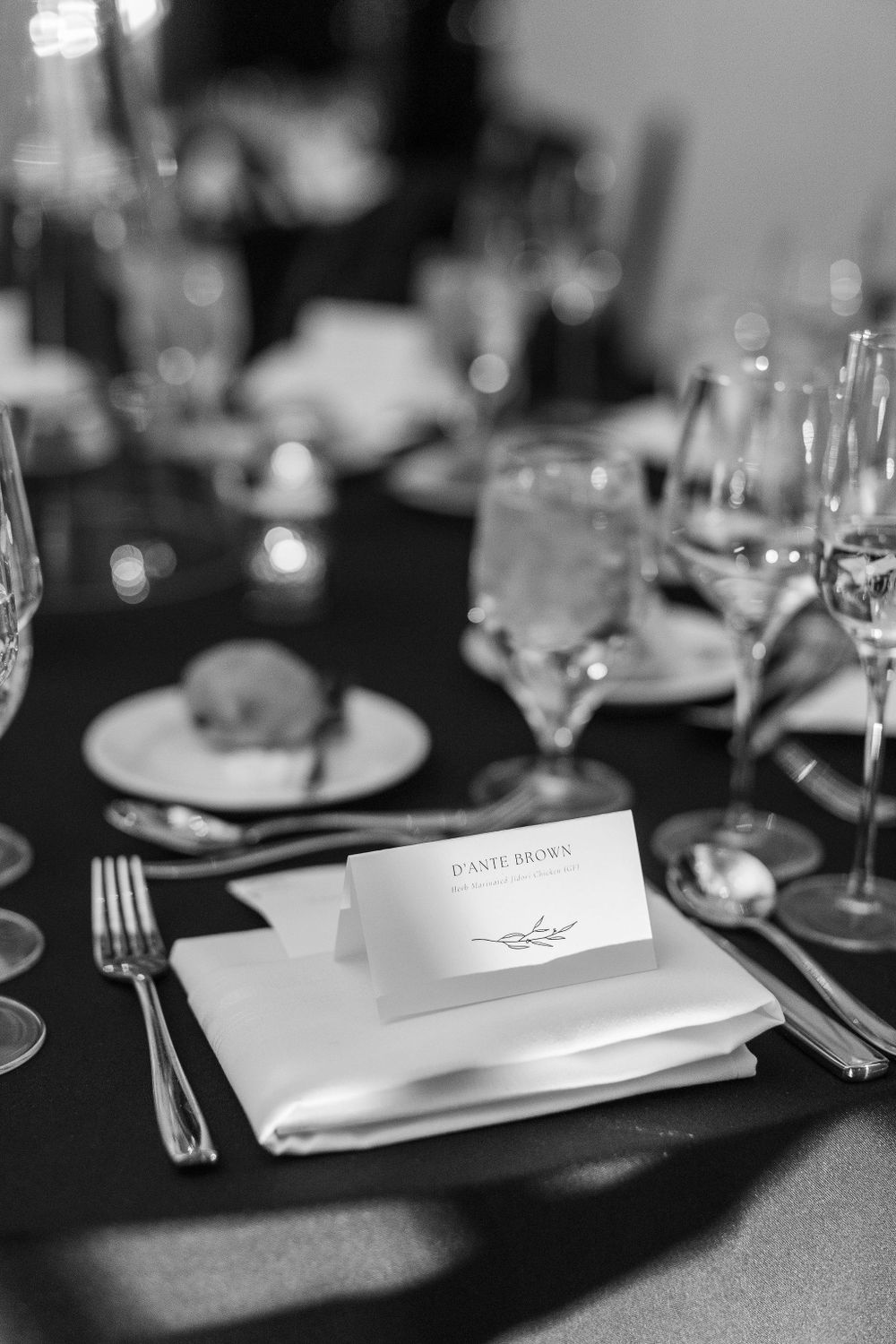 An elegant place setting with white napkin and place card on a dark table at a formal wedding reception.