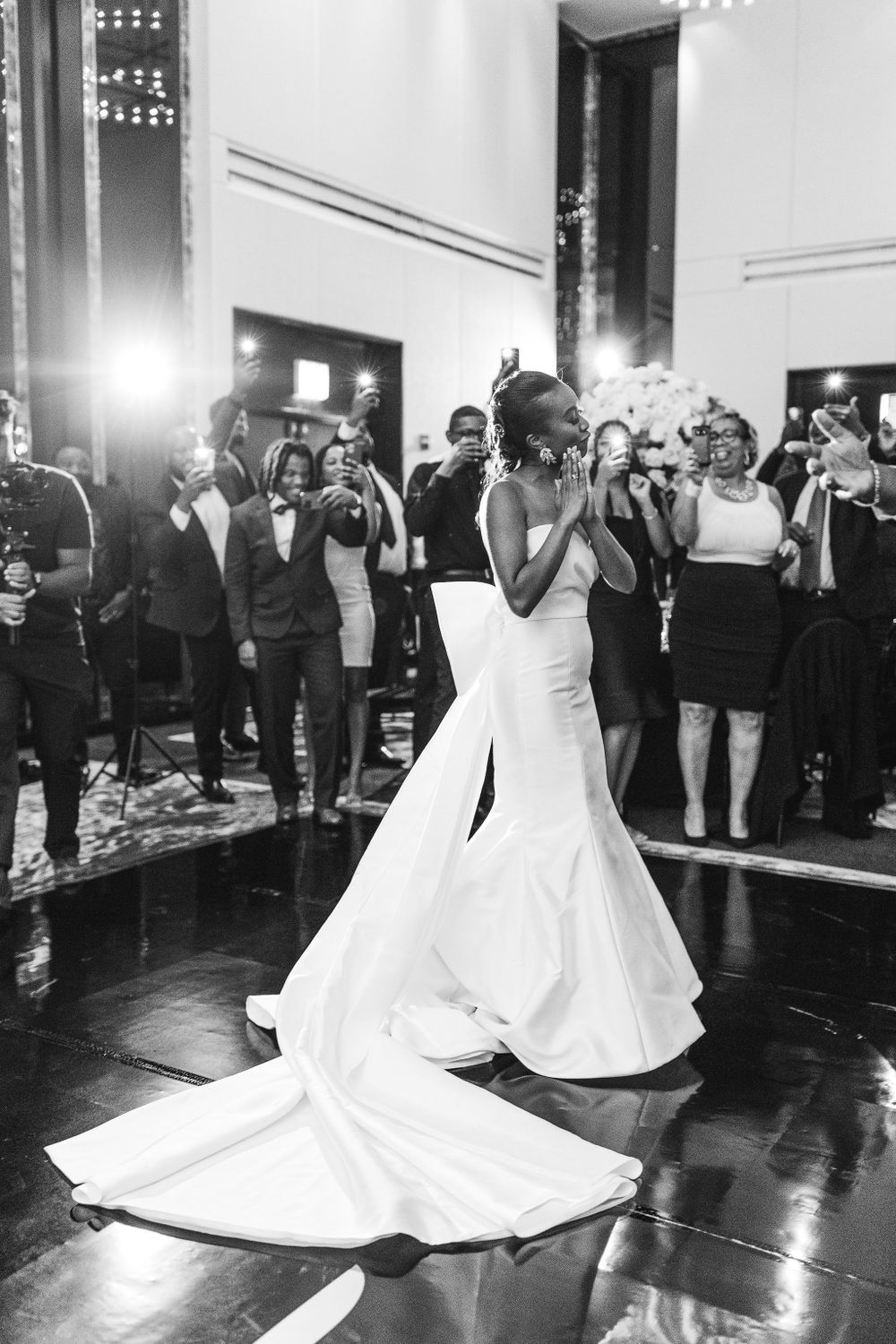 A stunning bride in a white mermaid wedding gown dances on a polished floor while guests watch and take photos.
