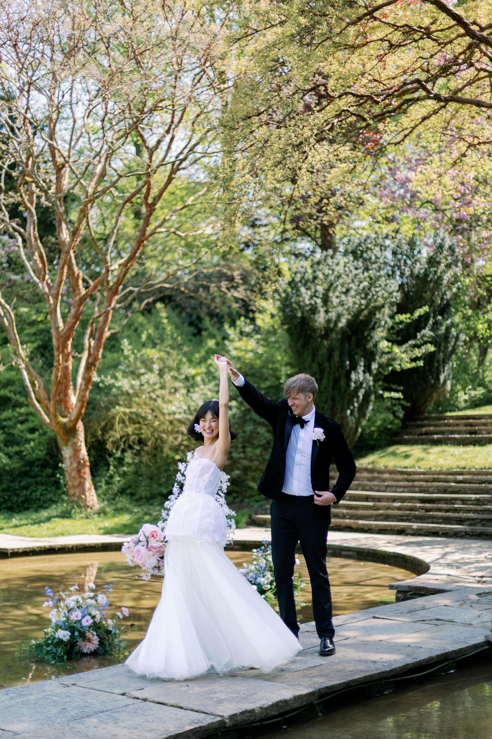 A joyful wedding couple poses together by a garden fountain surrounded by blooming trees and stone steps.