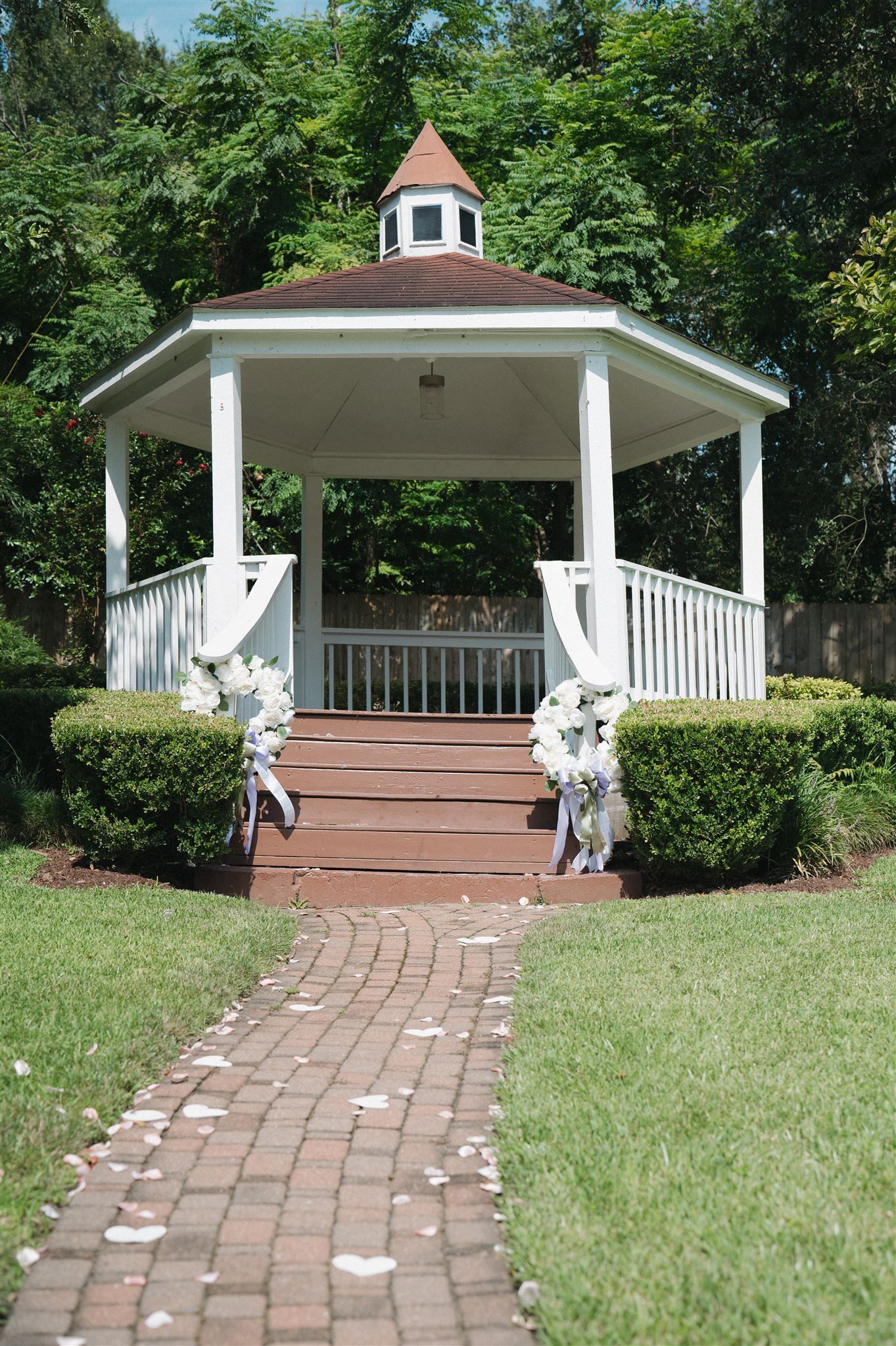 A white wedding gazebo with floral decorations and a curved brick pathway leading to wooden steps through a manicured lawn.