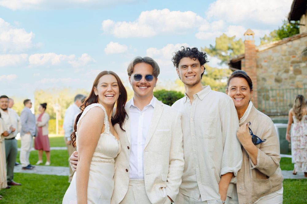 Wedding guests gather on a sunny lawn outside a rustic stone villa wearing light summer attire.