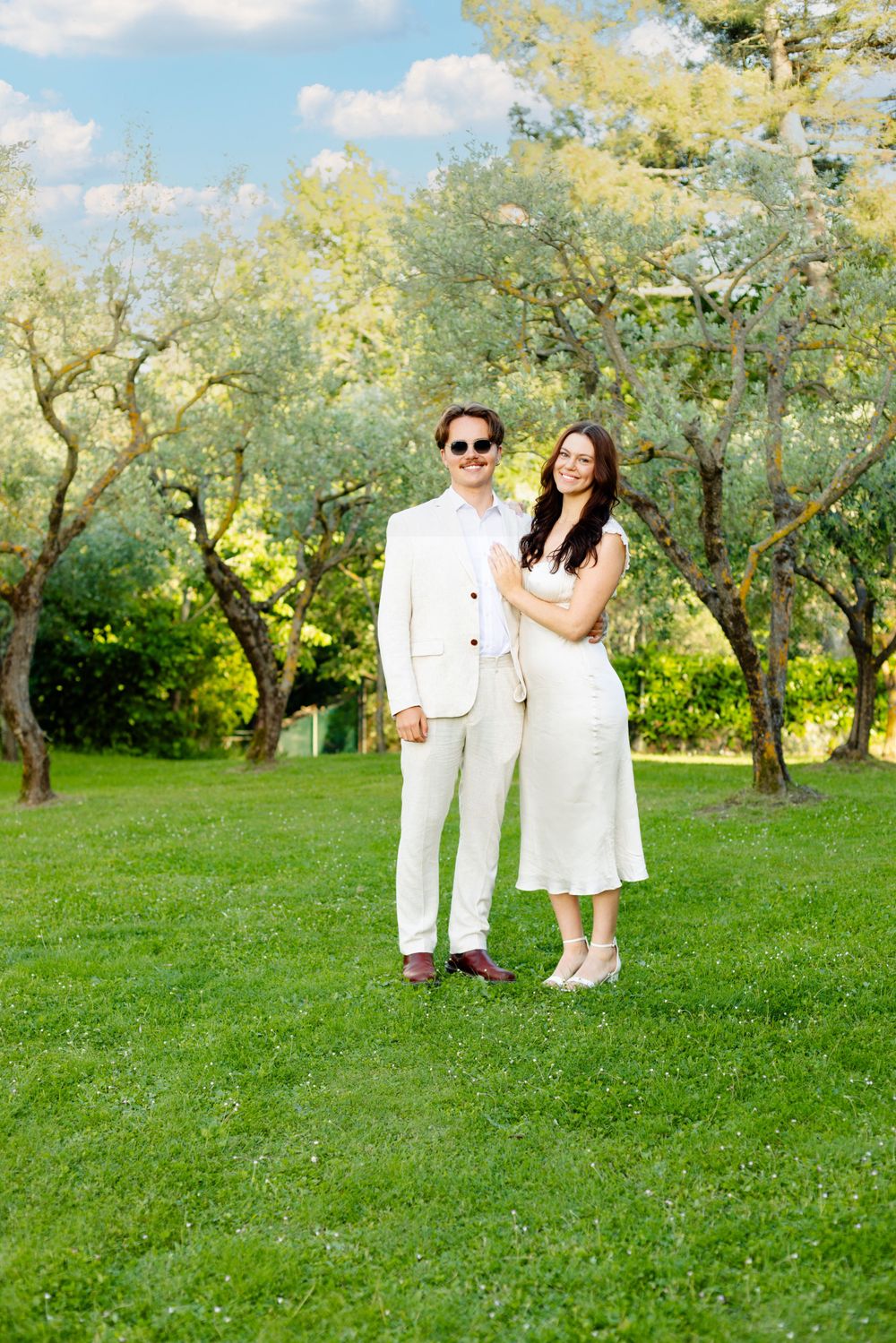 A couple in white outfits poses together on lush green grass in an olive grove.