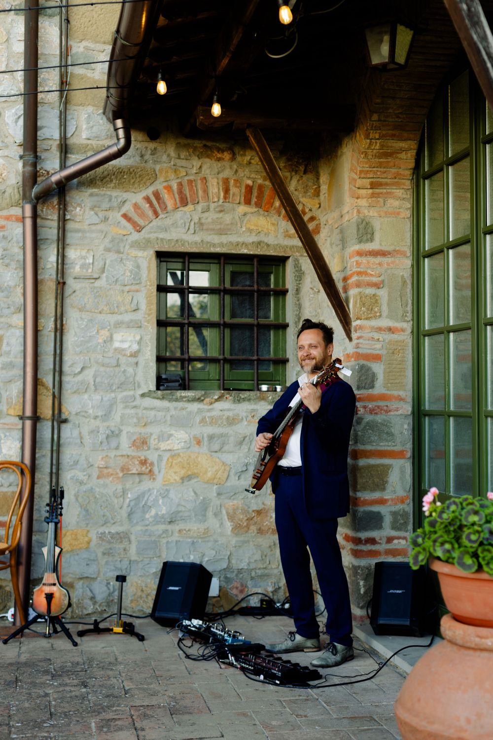 Musician performing with a violin against rustic stone wall with string lights overhead.
