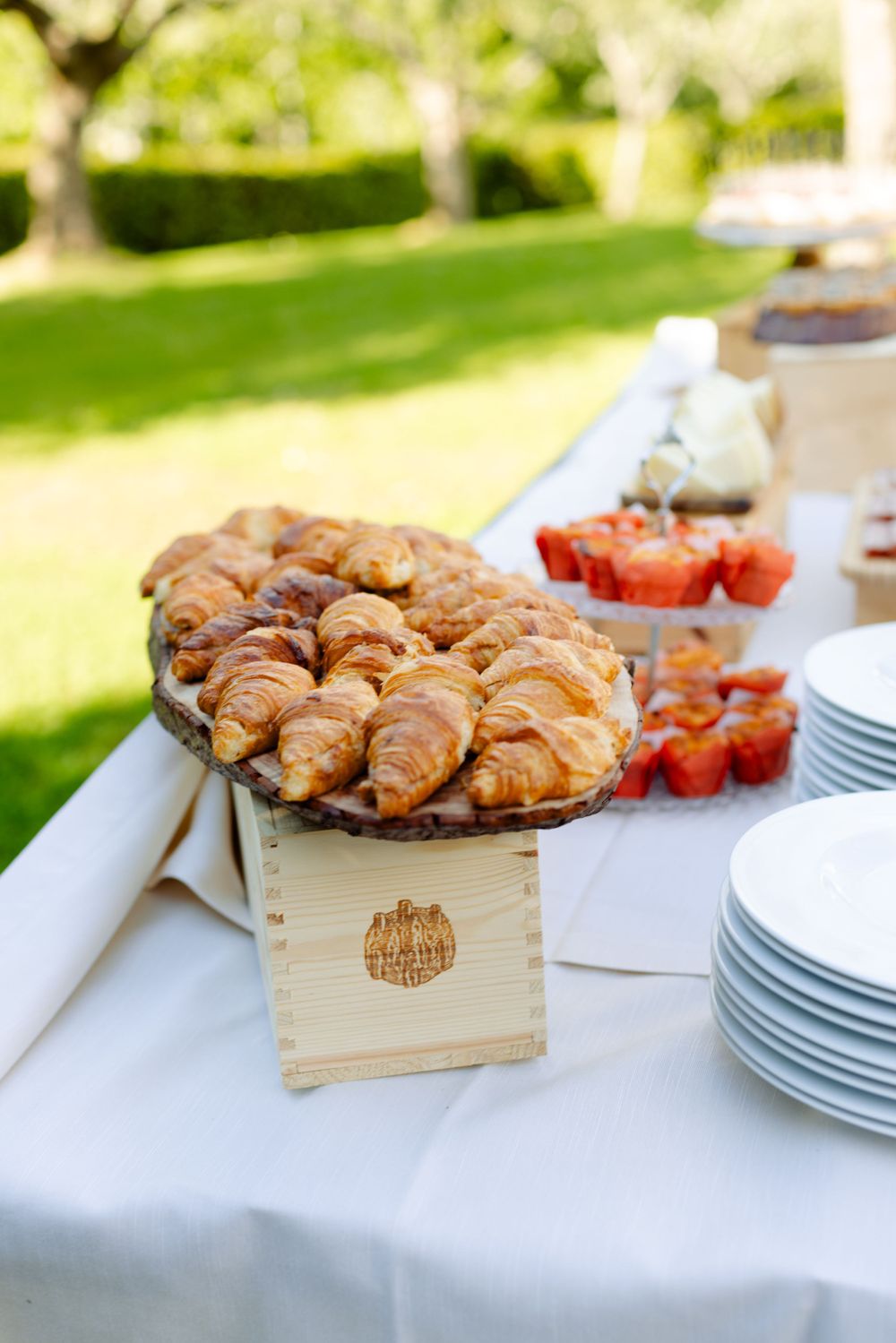 Fresh croissants and pastries displayed on wooden box at outdoor brunch setup.