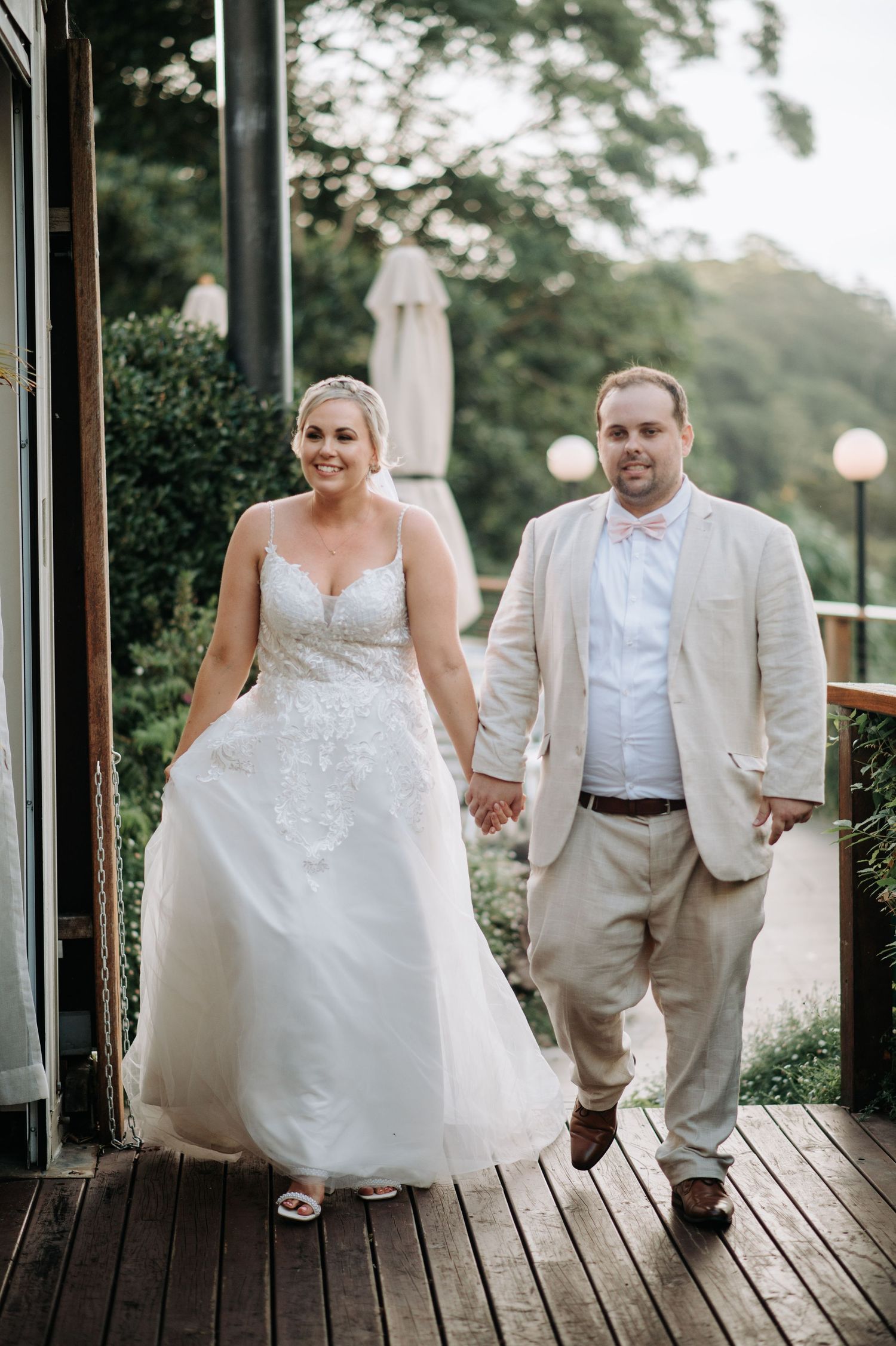 Newlyweds walk together on wooden deck surrounded by greenery.