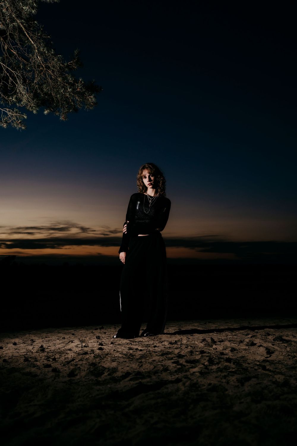 A silhouetted figure stands on sandy ground against a dramatic sunset sky with deep blue tones.