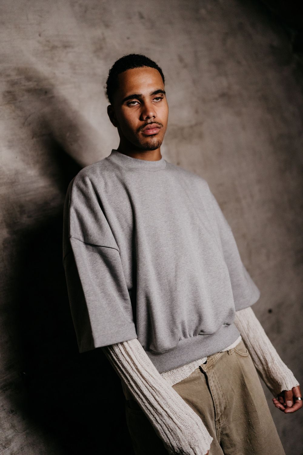 A person wearing a gray sweatshirt and beige pants poses against a dark textured background in a fashion editorial style.