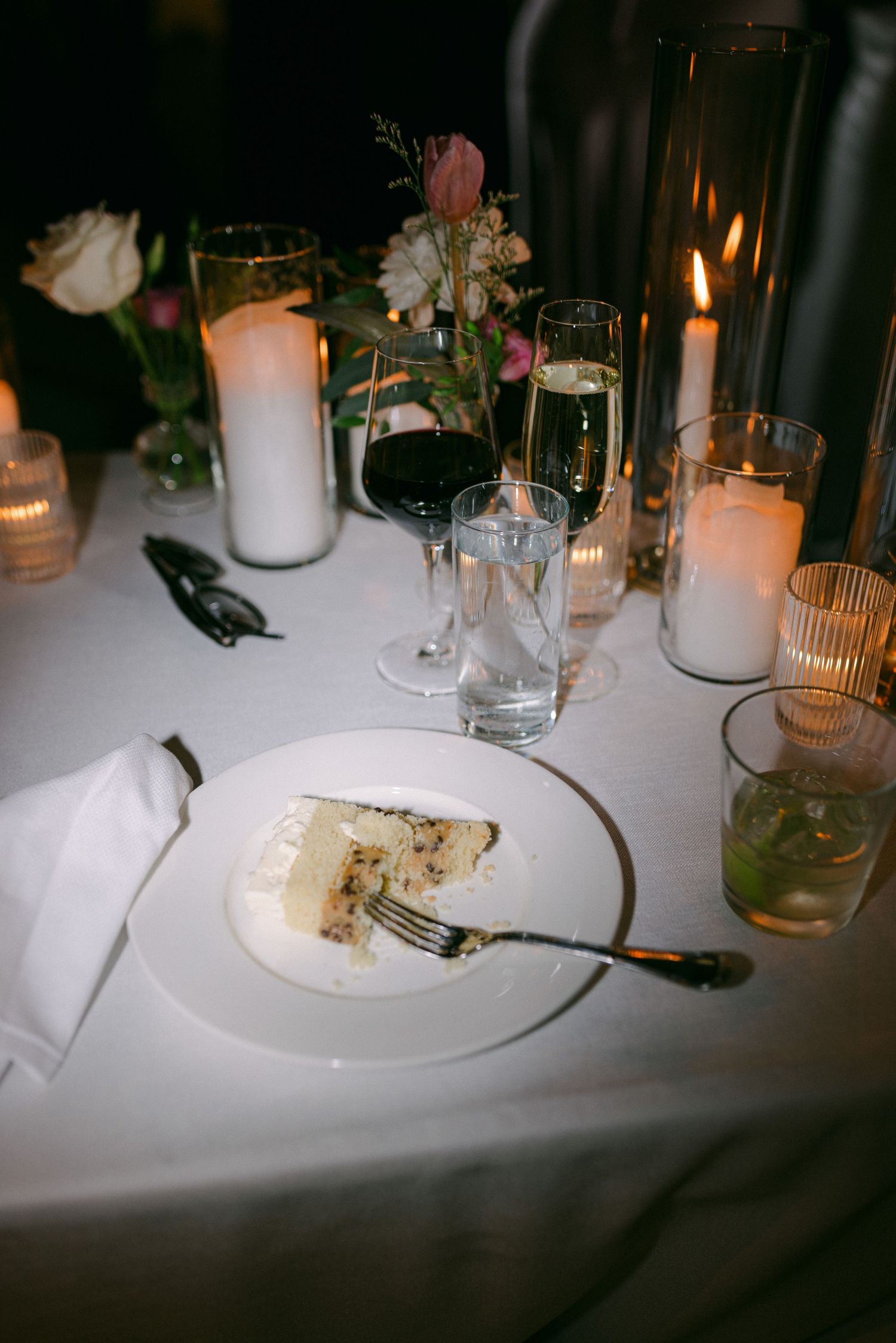 Wedding reception table setting with candlelight, wine glasses, and partially eaten cake on white tablecloth.