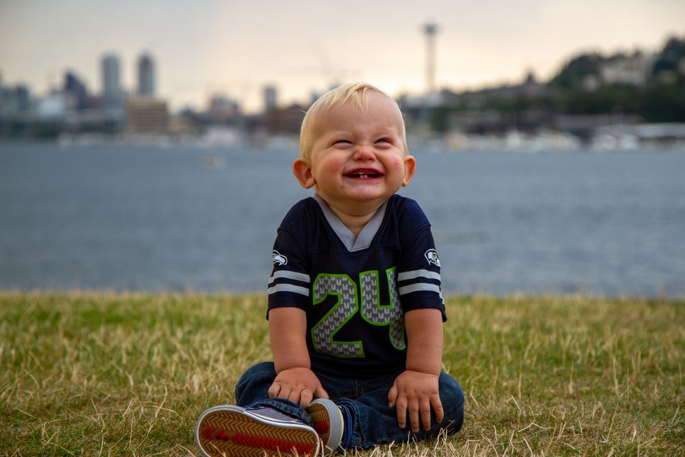 Cheerful toddler in navy jersey sits on grass with Seattle skyline in background.