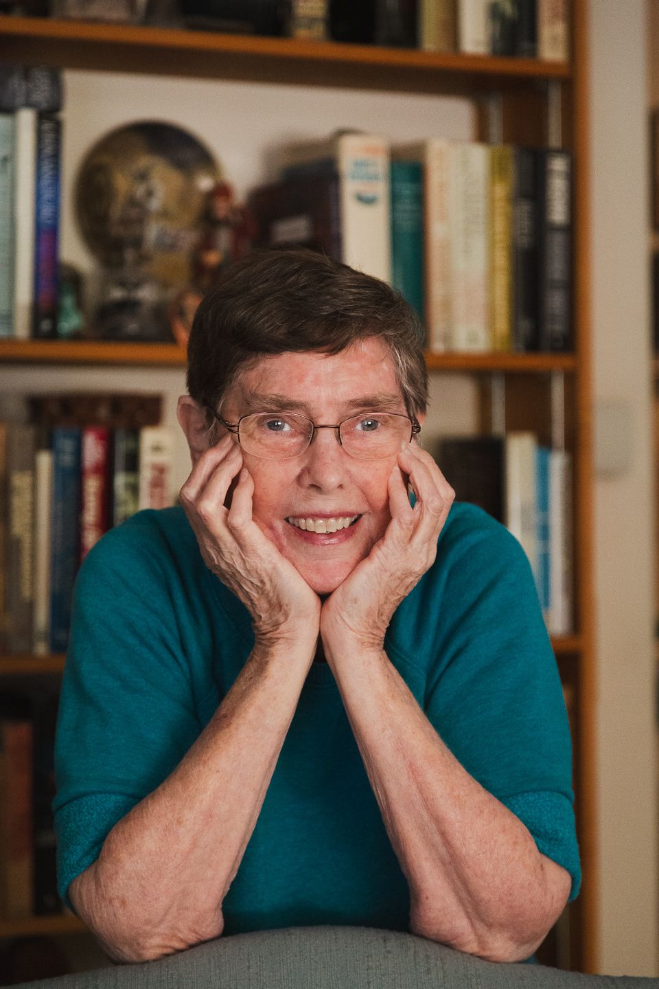 Senior in teal sweater smiling with hands on cheeks in front of bookshelf.