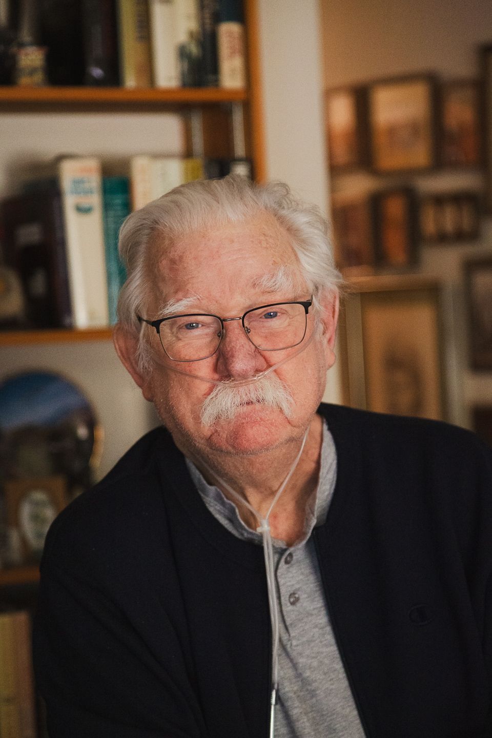 Senior in glasses and grey sweater sitting in front of bookshelf at home.