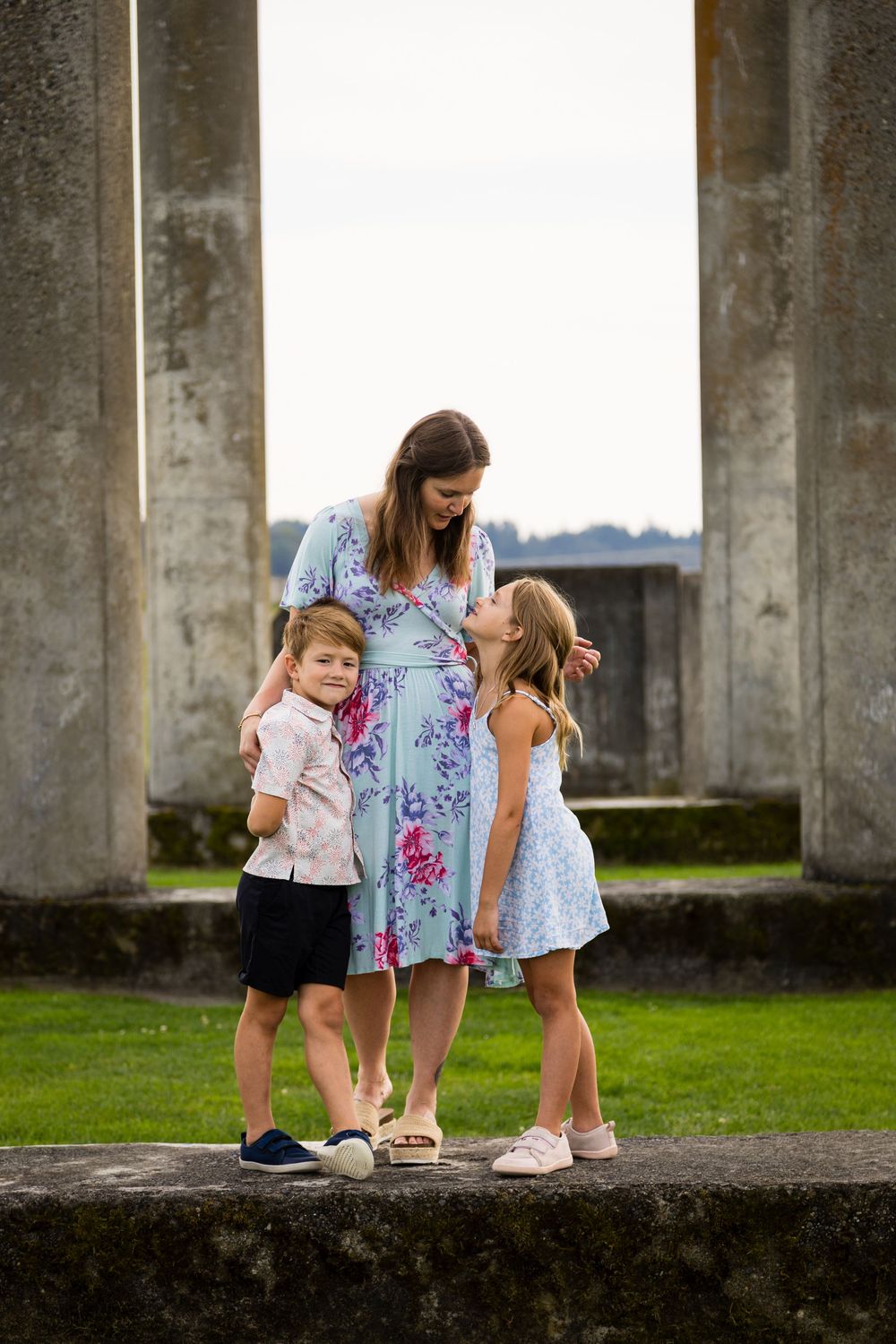 A family group interacts playfully among concrete pillars in an outdoor setting with green grass.