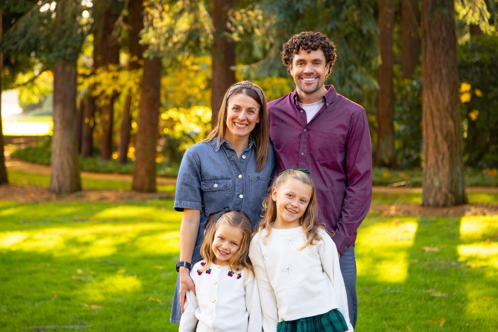 A family poses together outdoors in a sunlit wooded park setting during a photo session.