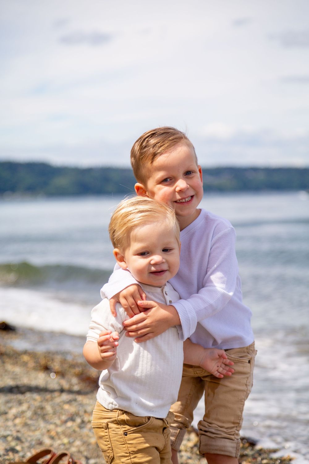 Two children embrace and smile while standing on a rocky beach with blue water and mountains in the background.