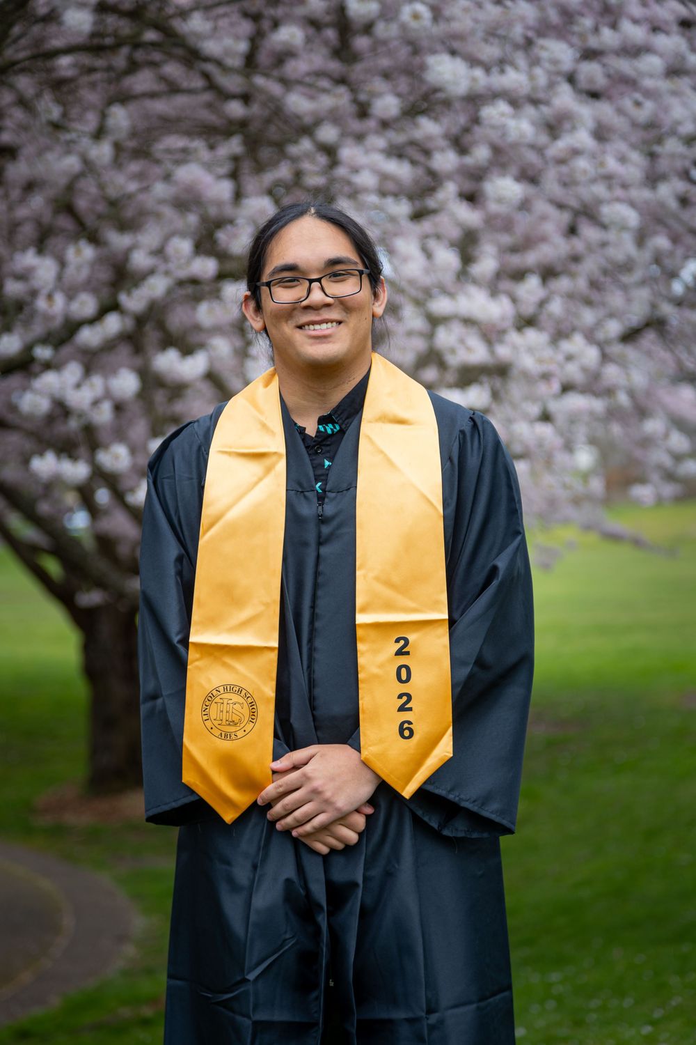 Smiling graduate in black gown and gold 2026 stole poses outdoors near blooming cherry blossom trees.