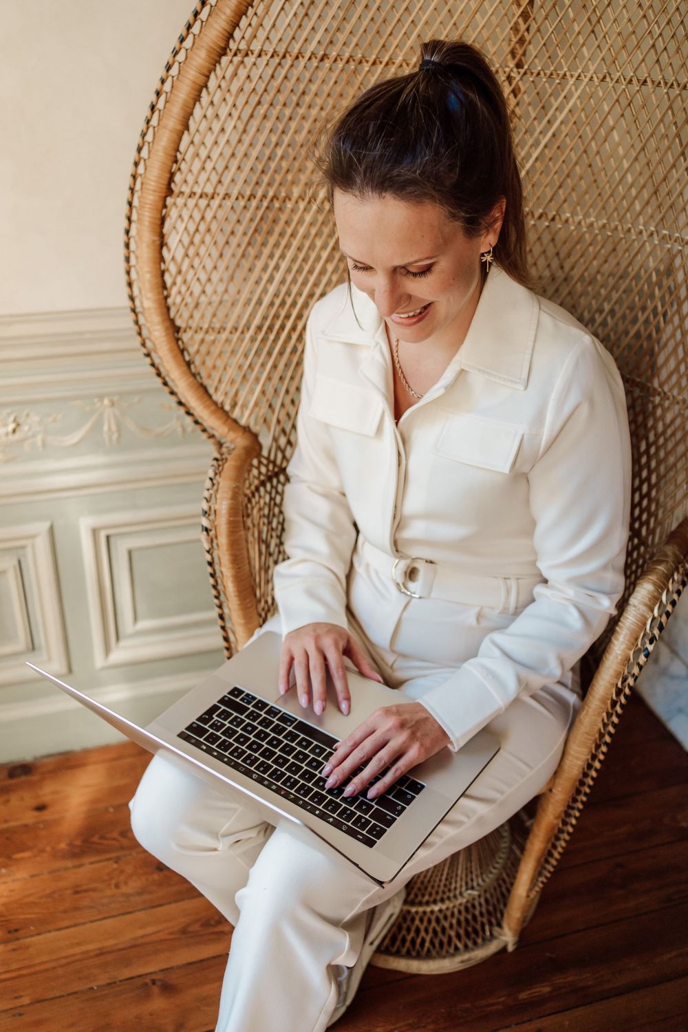 vrouw met beige jumpsuit zittende in rotanstoel met laptop op de schoot