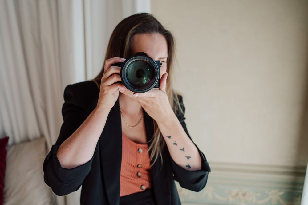 vrouw met zwarte blazer en oranje t-shirt met fototoestel in de hand zittende op een kruk