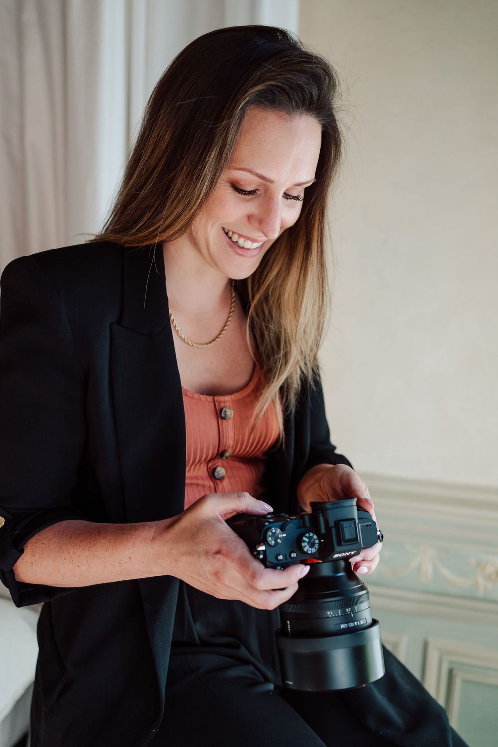 vrouw met zwarte blazer en oranje t-shirt met fototoestel in de hand zittende op een kruk