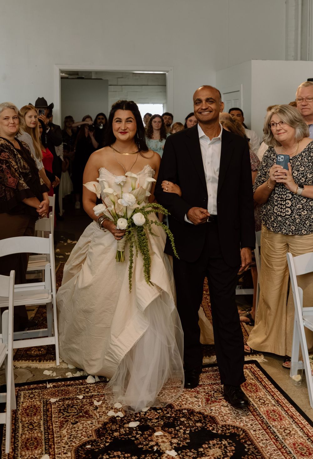 Wedding ceremony processional in indoor venue with oriental rug and white chairs.
