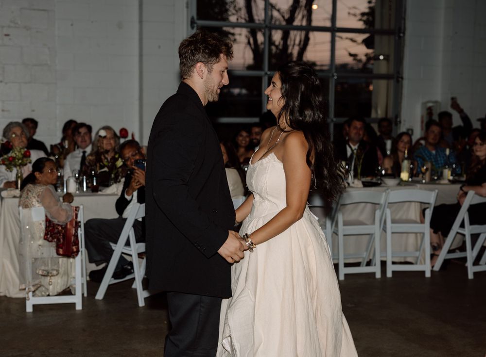 A couple shares their first dance at a wedding reception with guests seated at tables in the background.
