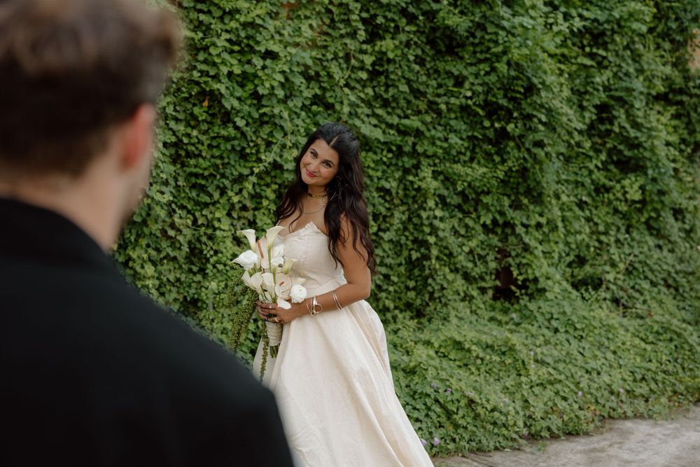 A bride in a white dress holds calla lilies against an ivy-covered wall.