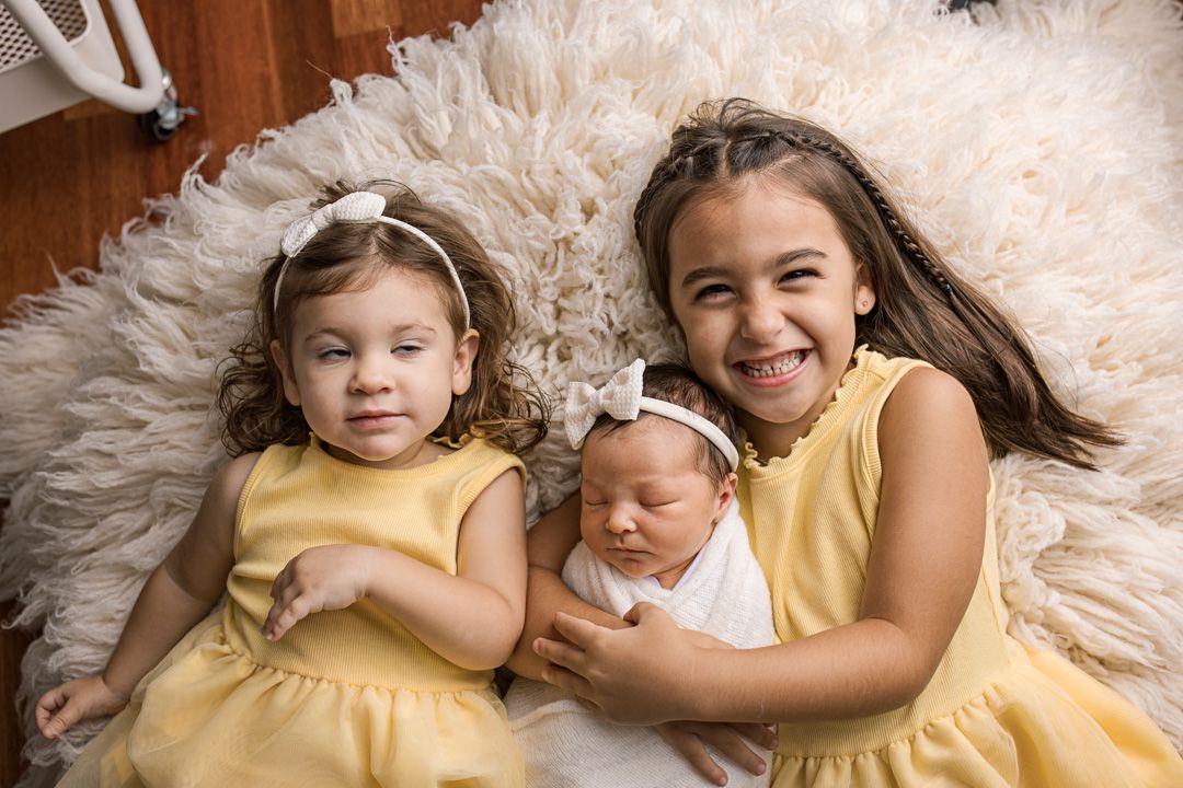 Three siblings in matching yellow dresses pose together on a fluffy white rug for a sweet family portrait.