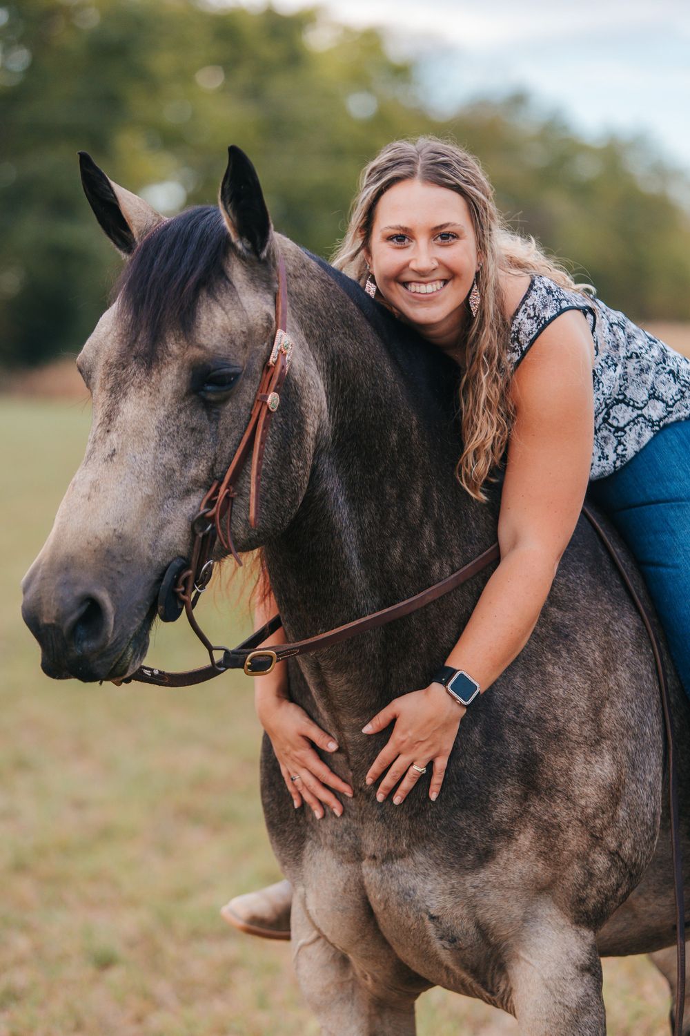 Grey horse with bridle being affectionately embraced in a grassy field.