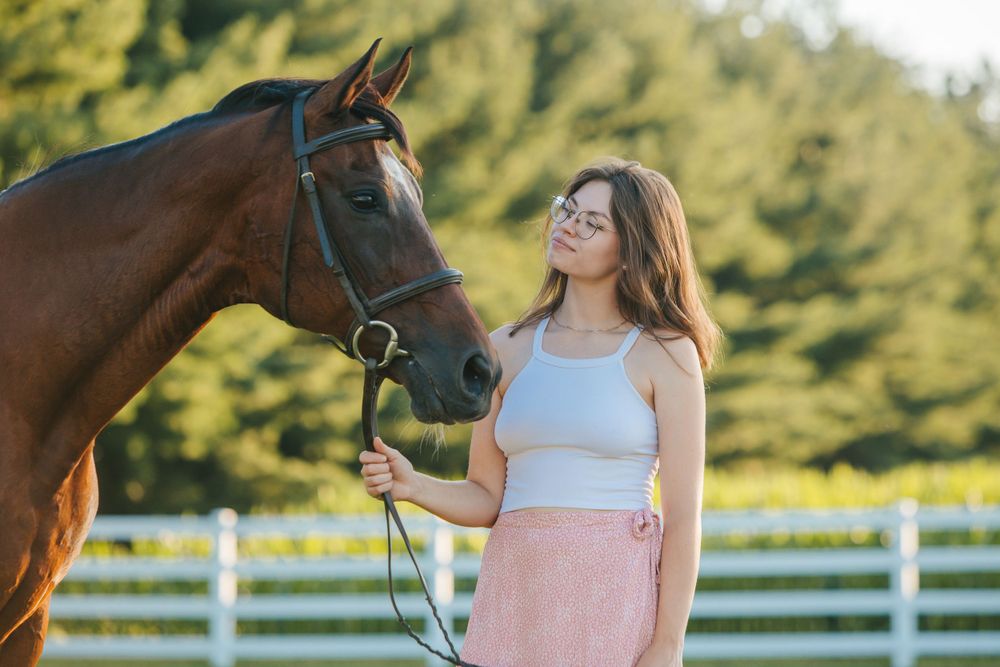 A bay horse and someone in a pink skirt share a tender moment in front of white fencing.