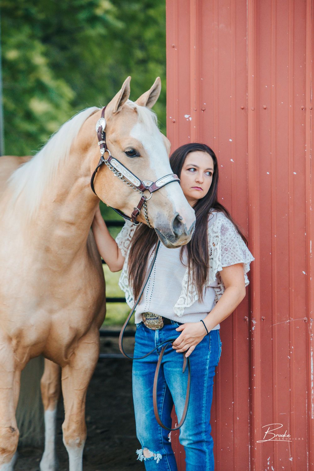 A palomino horse poses with someone in western attire against a red barn wall.
