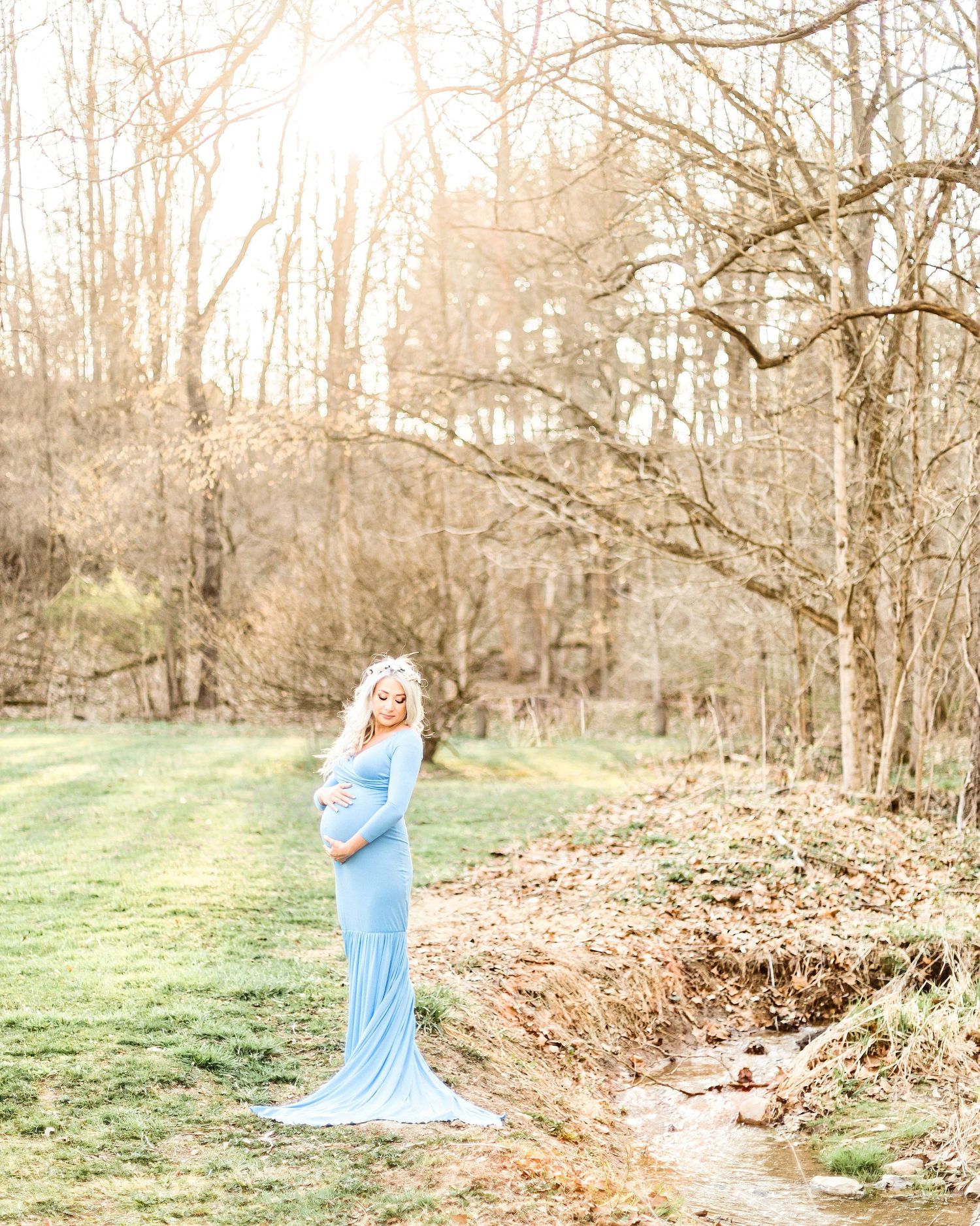A maternity photo of a woman in a flowing blue dress standing in a sunlit wooded area. Pittsburgh maternity photographer