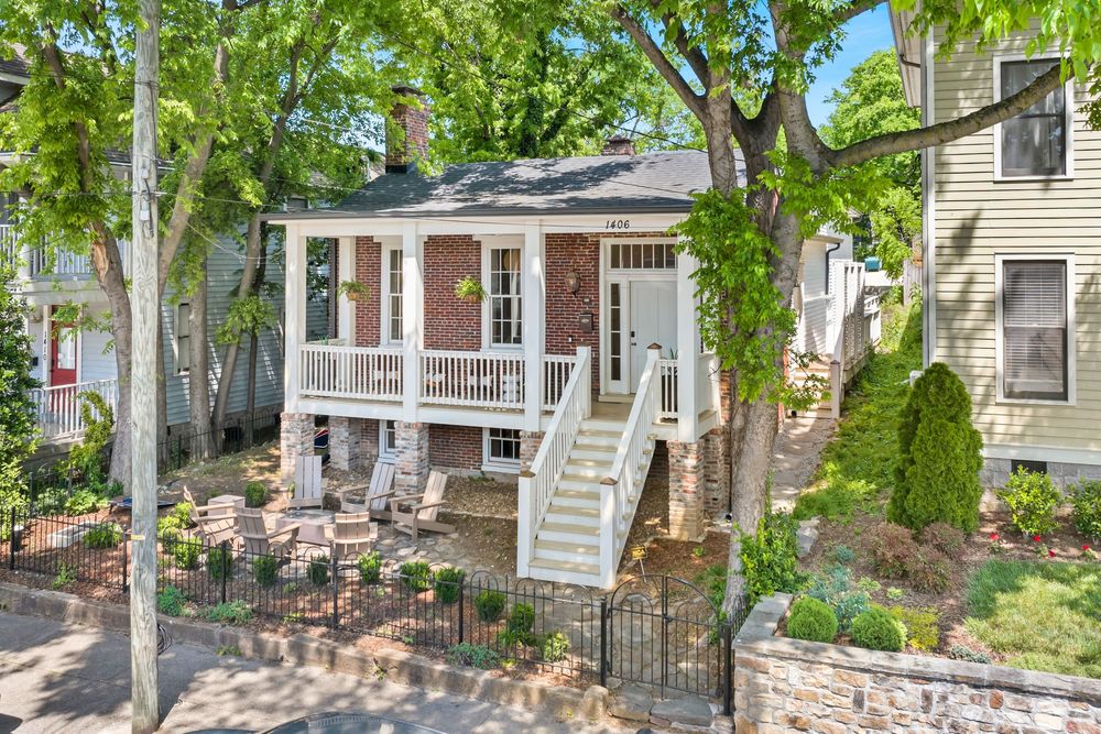 Charming raised cottage home with covered front porch, brick facade, white railings, and lush mature trees in a residential neighborhood.