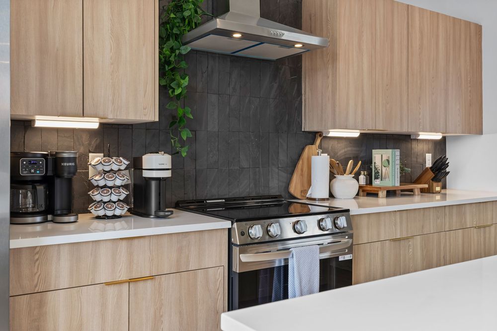 Modern kitchen with light wood cabinets, stainless steel range, dark tile backsplash, and under-cabinet lighting.