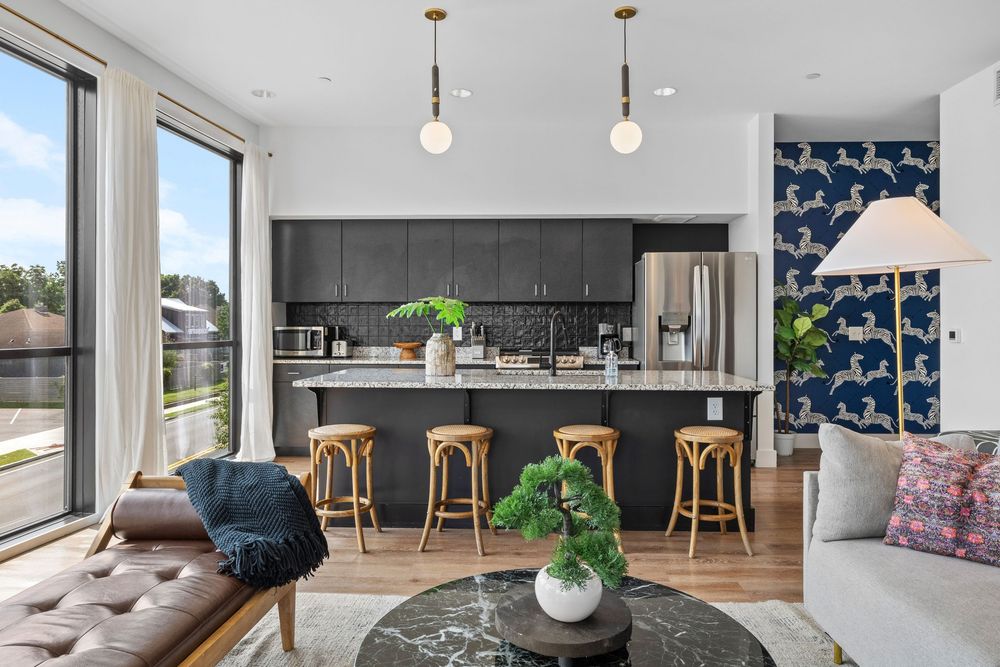 Modern open-concept kitchen and living room with dark cabinets, gold pendant lights, bar stools, and bold blue floral wallpaper.