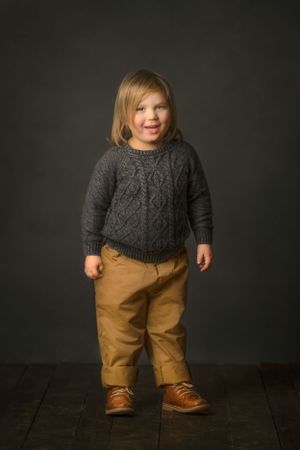 A child wearing a sophisticated gray cable knit sweater and khaki pants stands against a dark background.