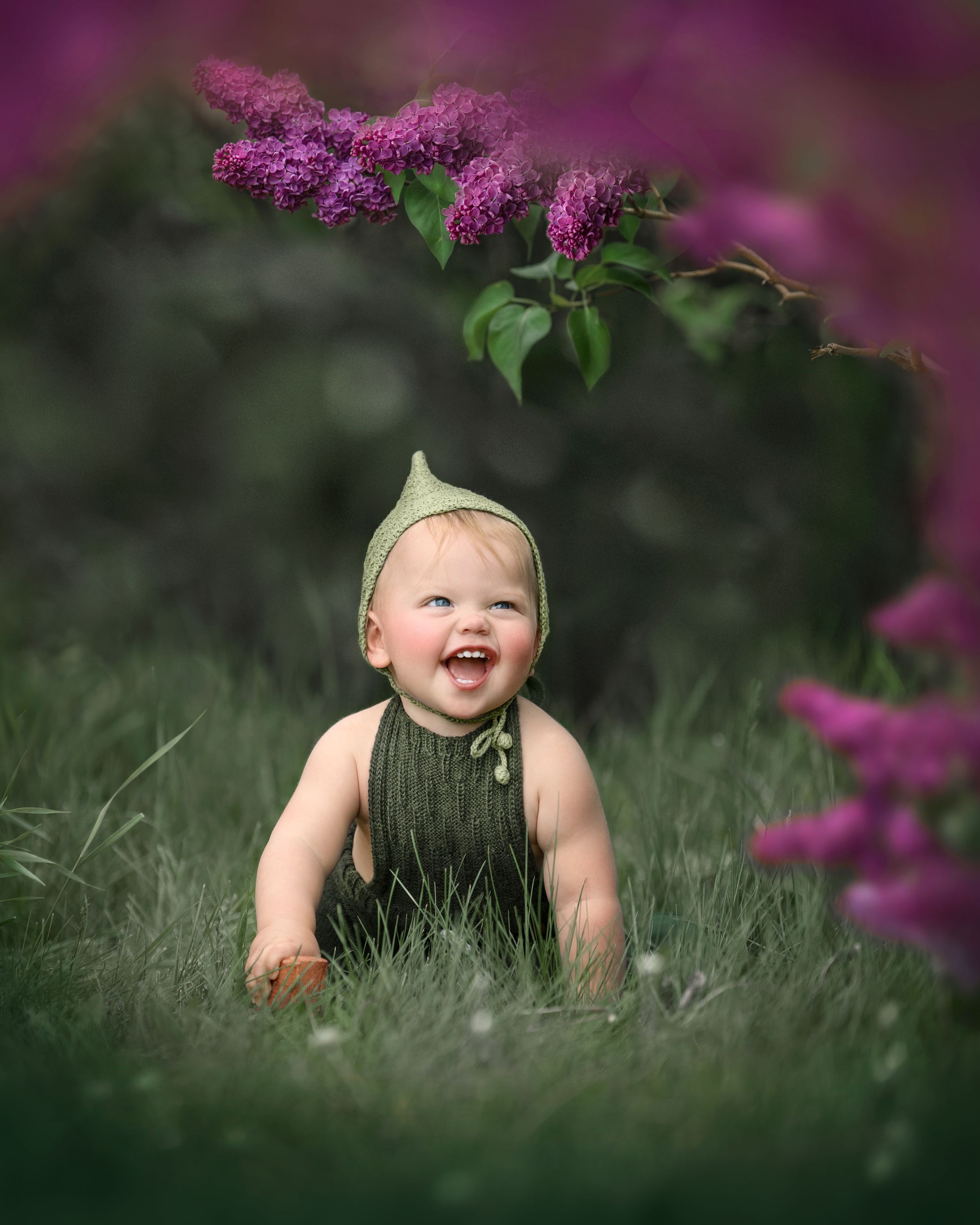 Baby in green outfit laughing under blooming lilac branches.