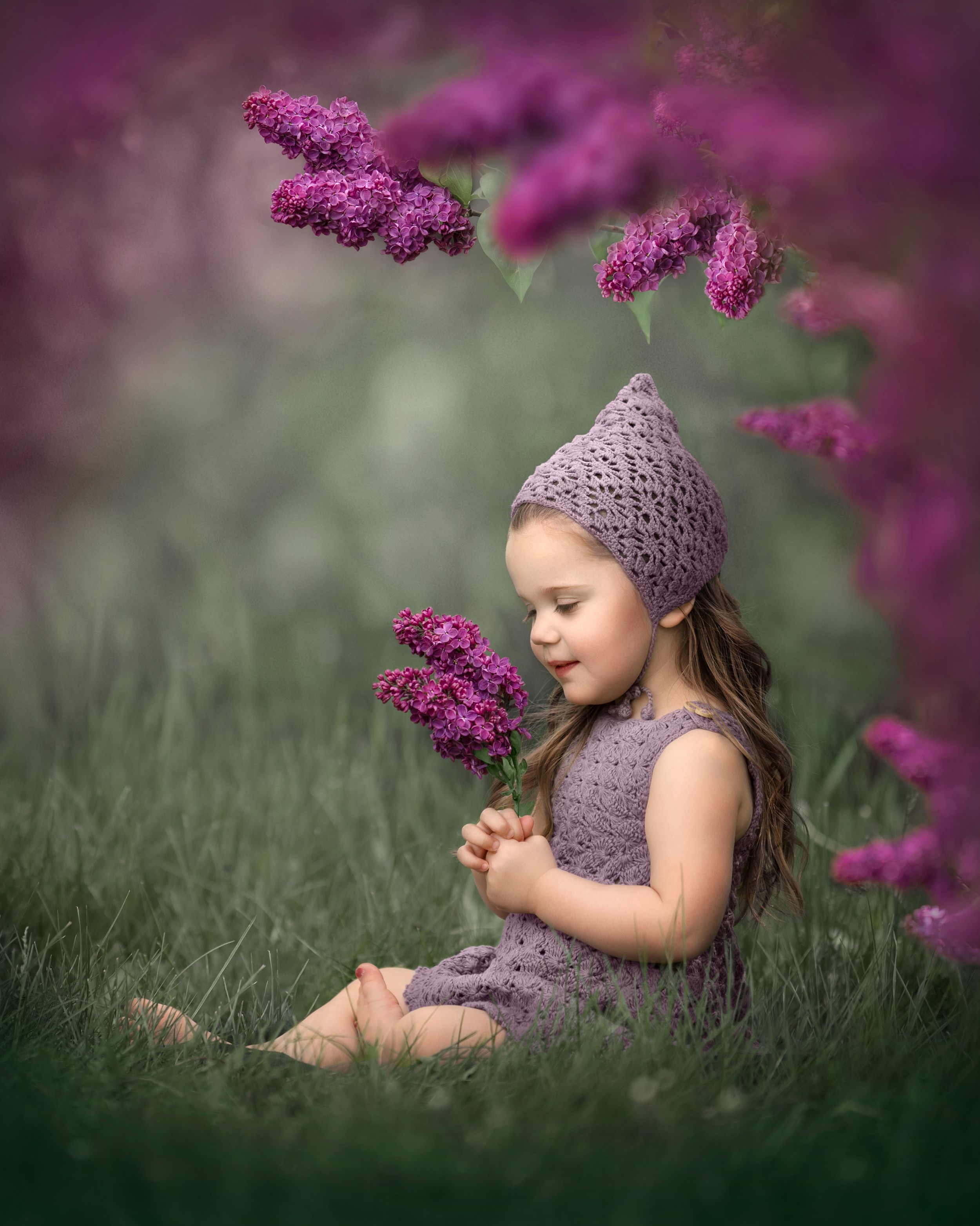 Child in purple dress sitting peacefully under flowering lilac branches.