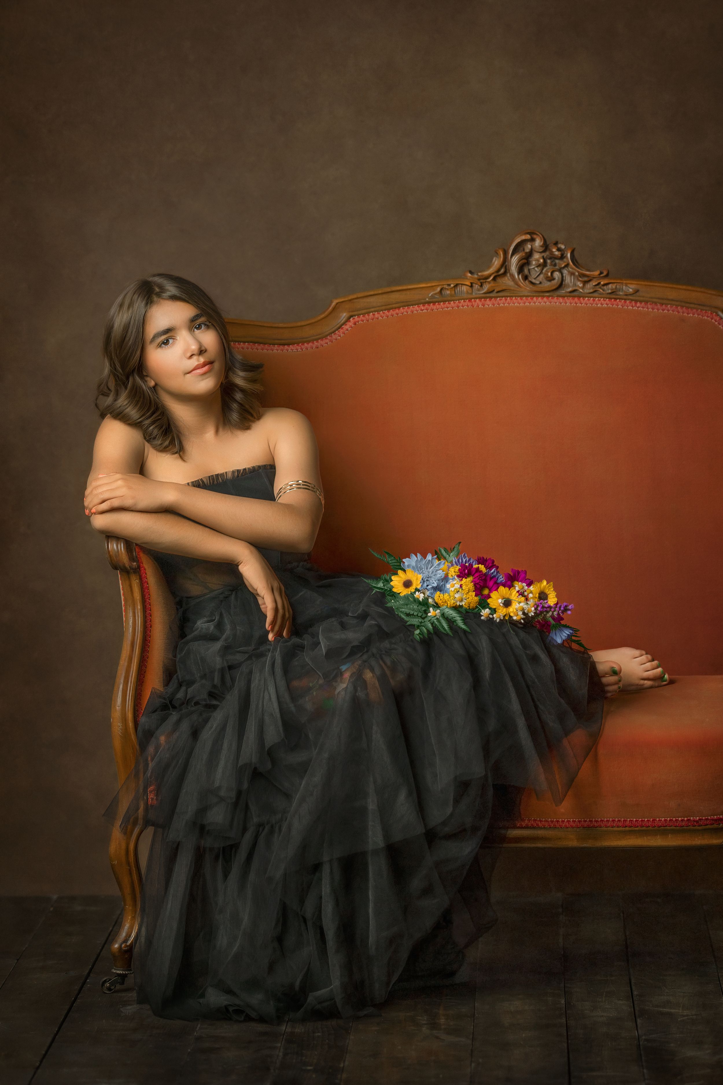 A dramatic portrait of a teen girl on an ornate orange vintage sofa with cheerful, colorful flowers and a flowing black formal gown.
