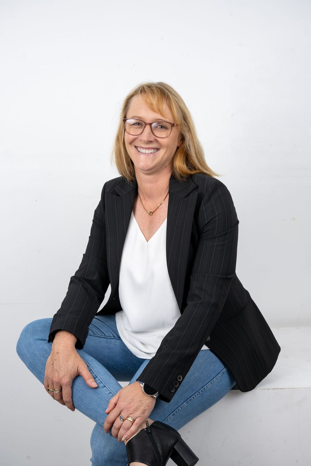 Smiling middle-aged woman with glasses wearing a black blazer and jeans, sitting casually against a white background.