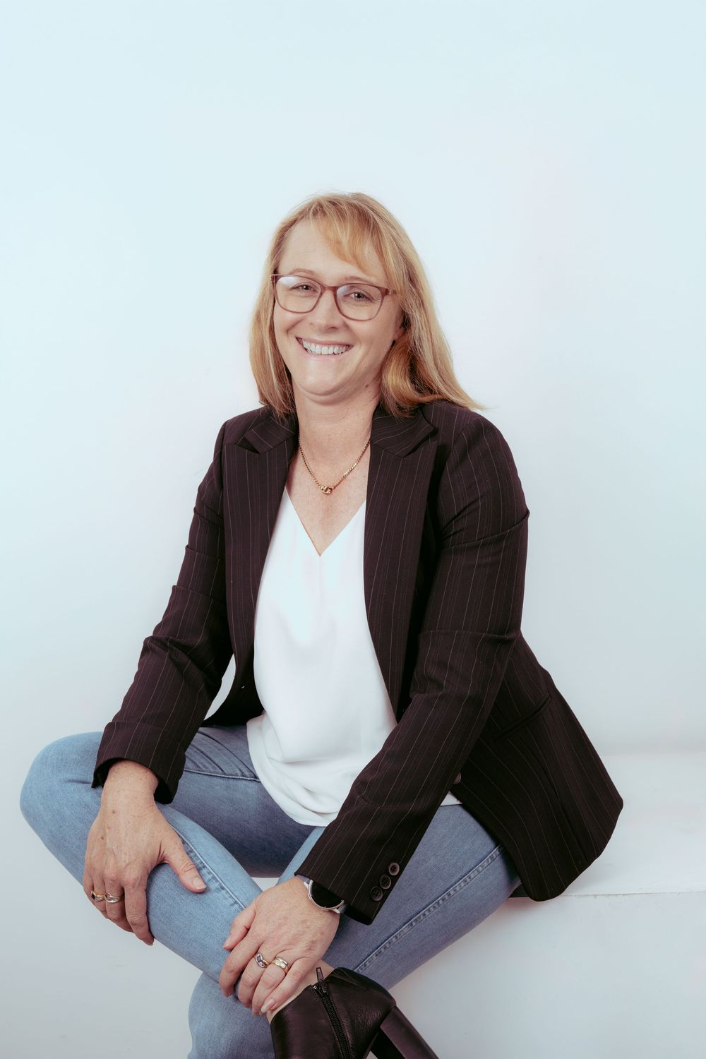 Smiling woman with glasses and blonde hair wearing a black blazer and jeans sitting casually against a white background.