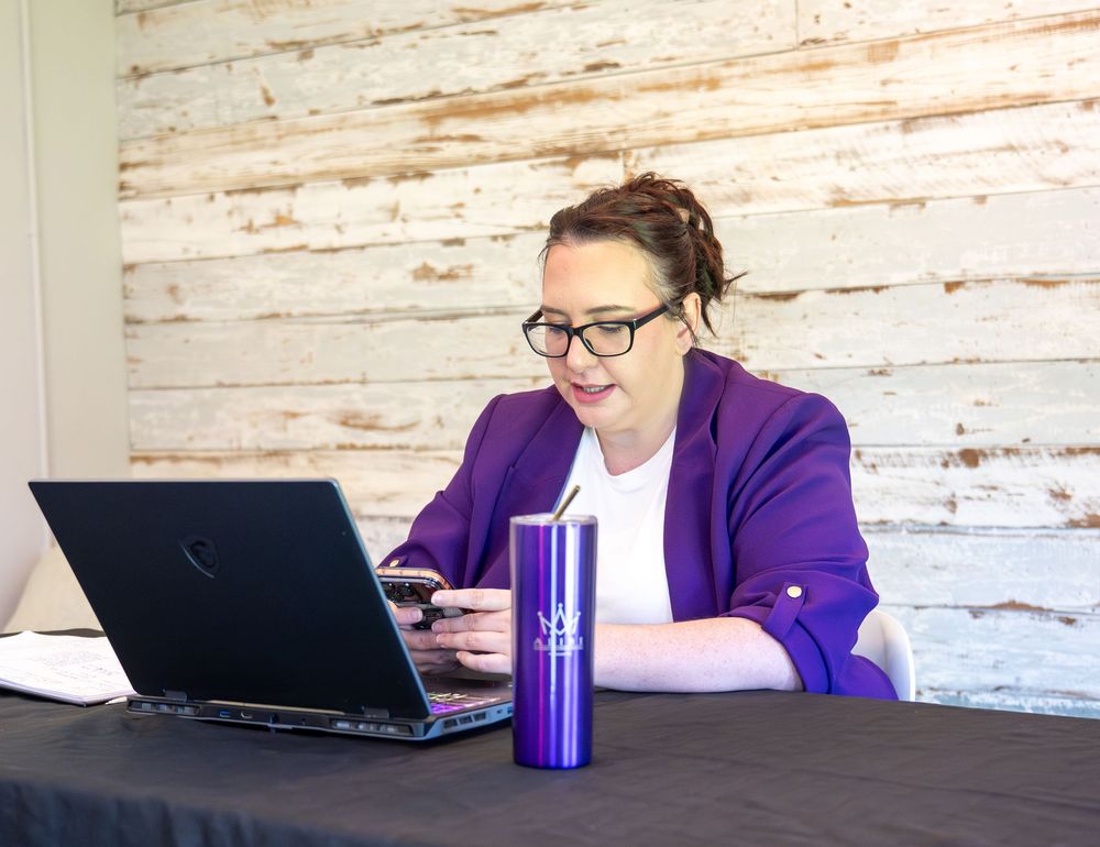 Woman in purple blazer smiling at laptop with blue tumbler on table against white wood wall.