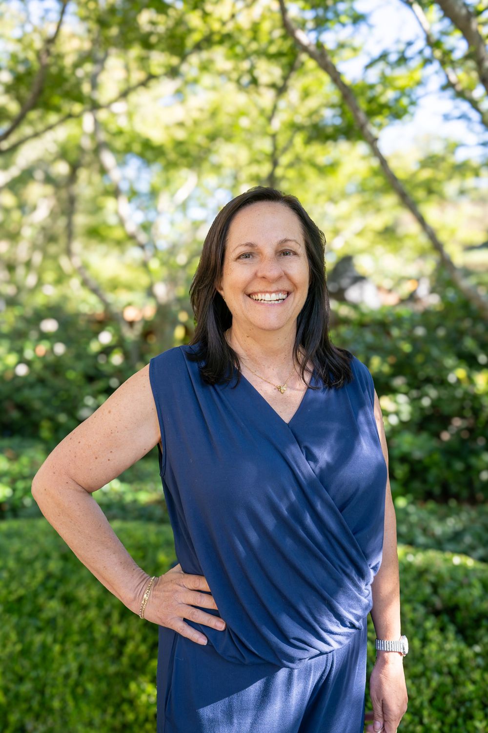 Smiling woman in blue sleeveless dress posing outdoors with lush green trees in background.