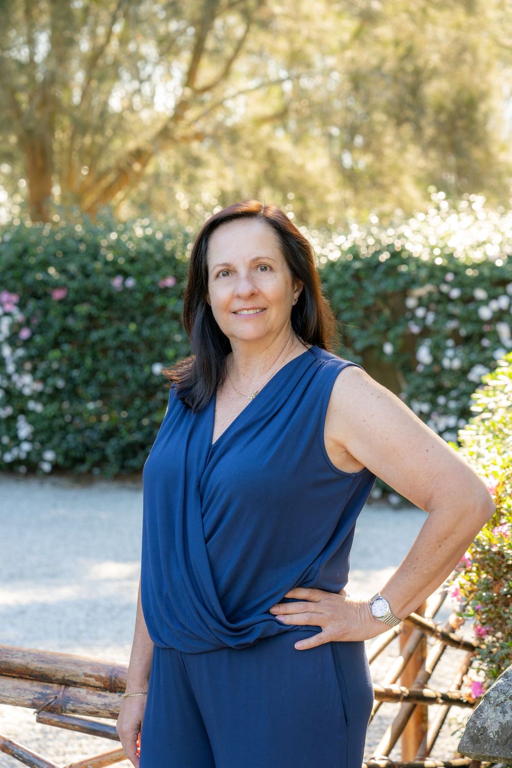 Woman in blue sleeveless dress posing outdoors in a garden with lush green hedges and pink flowers.