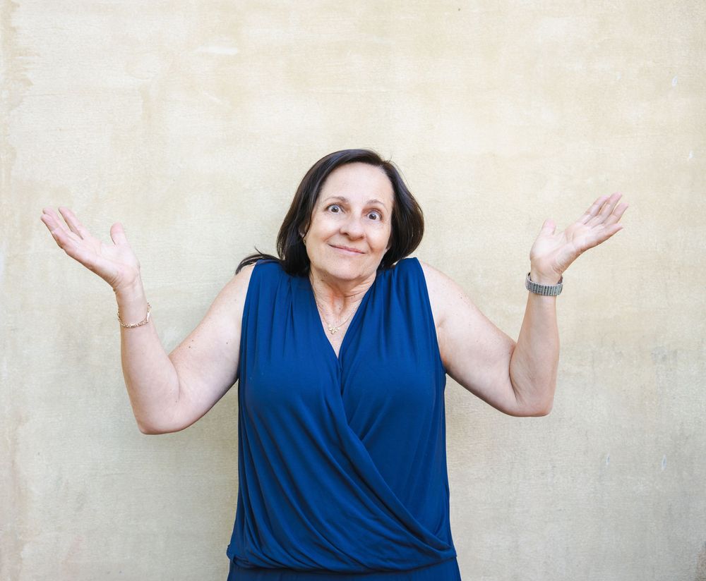 Middle-aged woman in blue sleeveless top shrugging with raised hands against beige wall.