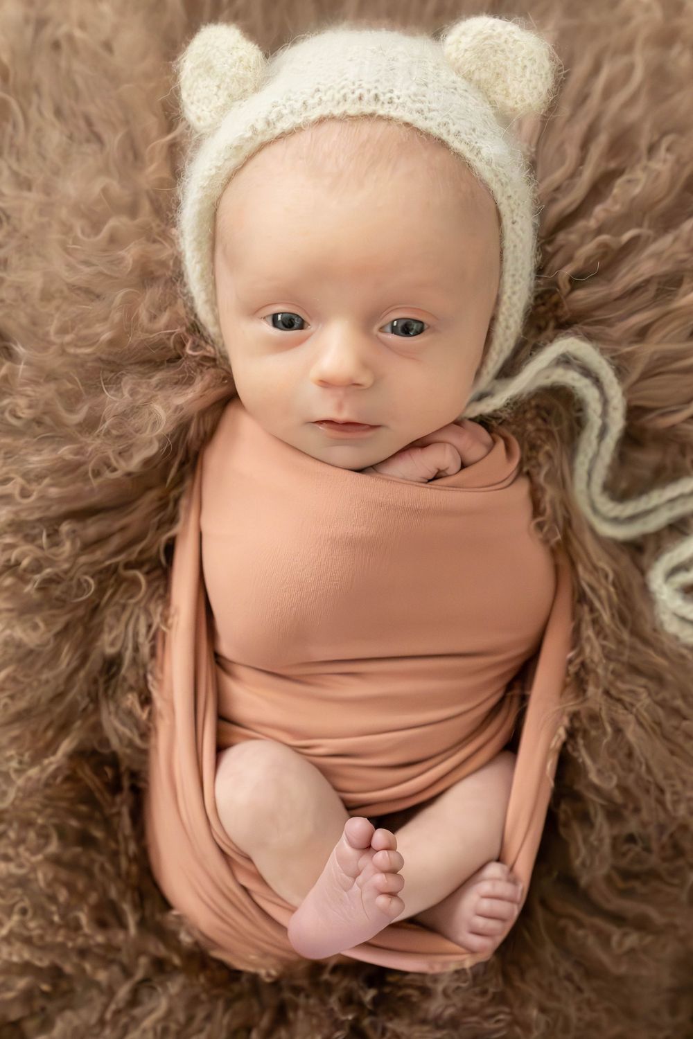 Sweet baby wearing knit bear hat and peach wrap lying on brown fur blanket during newborn photography session.