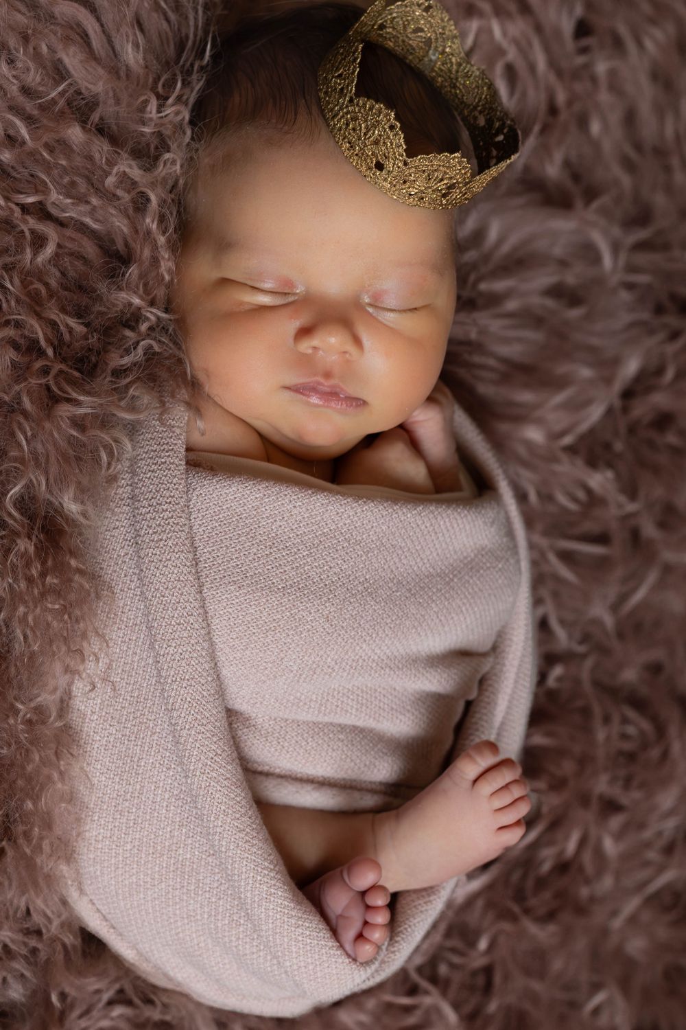 A sleeping newborn wrapped in a soft gray blanket wearing a delicate gold crown on a fluffy brown backdrop.