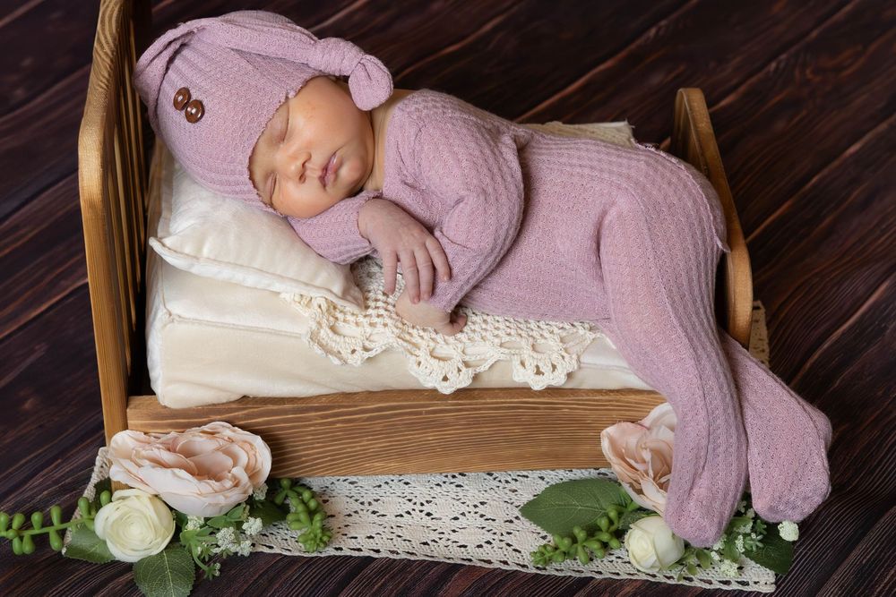 A sleeping baby wearing pink knit clothing lies on white lace fabric in a rustic wooden setting.