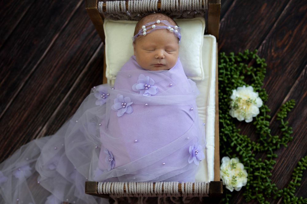 Sleeping newborn in purple tulle dress laying in rustic basket with white flowers.