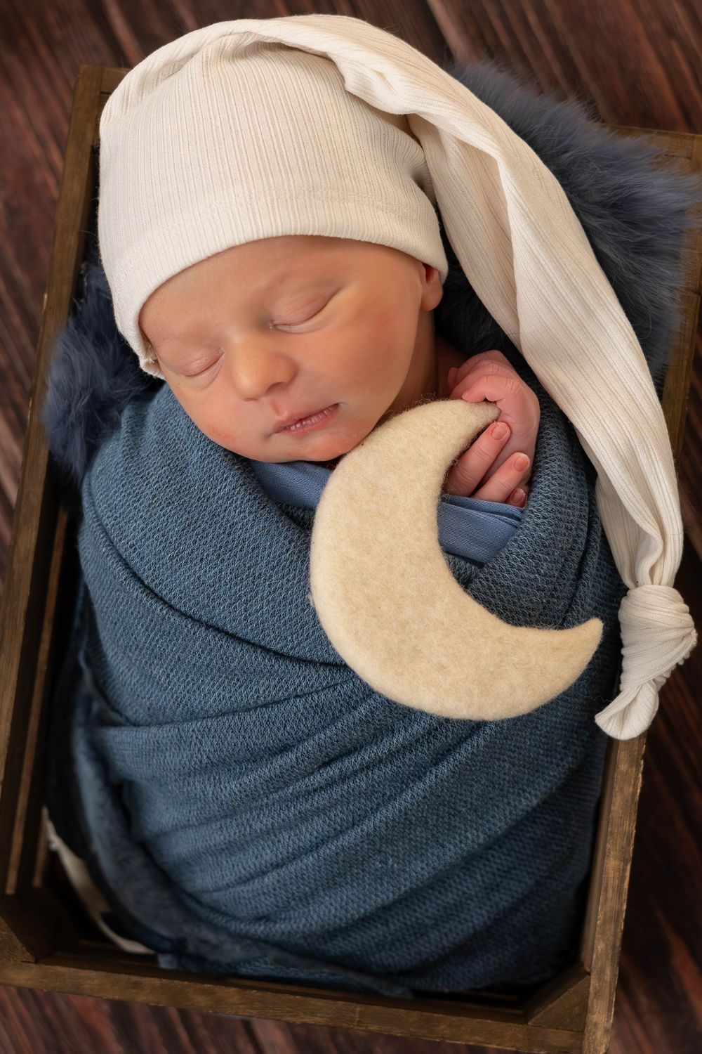 A sleeping newborn baby swaddled in blue fabric with a white hat and moon-shaped pillow prop in a wooden basket.