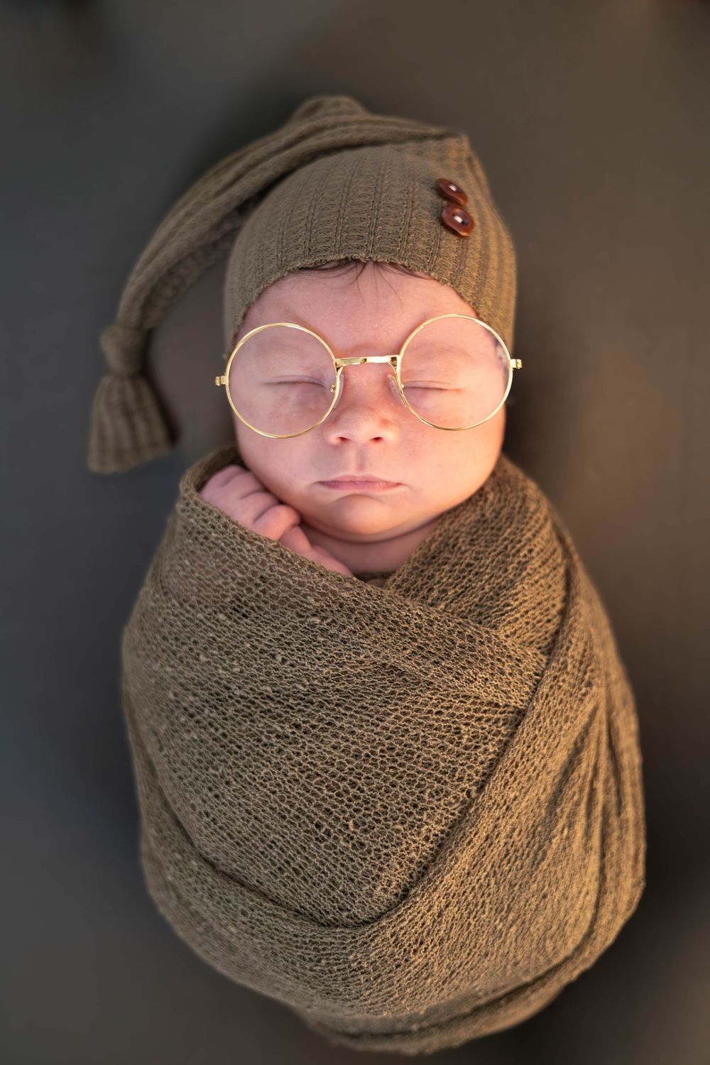 Sleeping newborn wrapped in brown knit blanket wearing round glasses and matching knit cap.